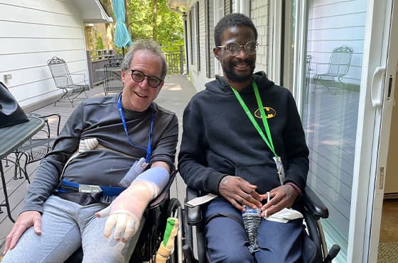 Two men in wheelchairs smiling on a sunlit balcony. One wears glasses and has a prosthetic hand, while the other has a Batman logo on his hoodie. Both have lanyards around their necks. Outdoor chairs and greenery are visible in the background.