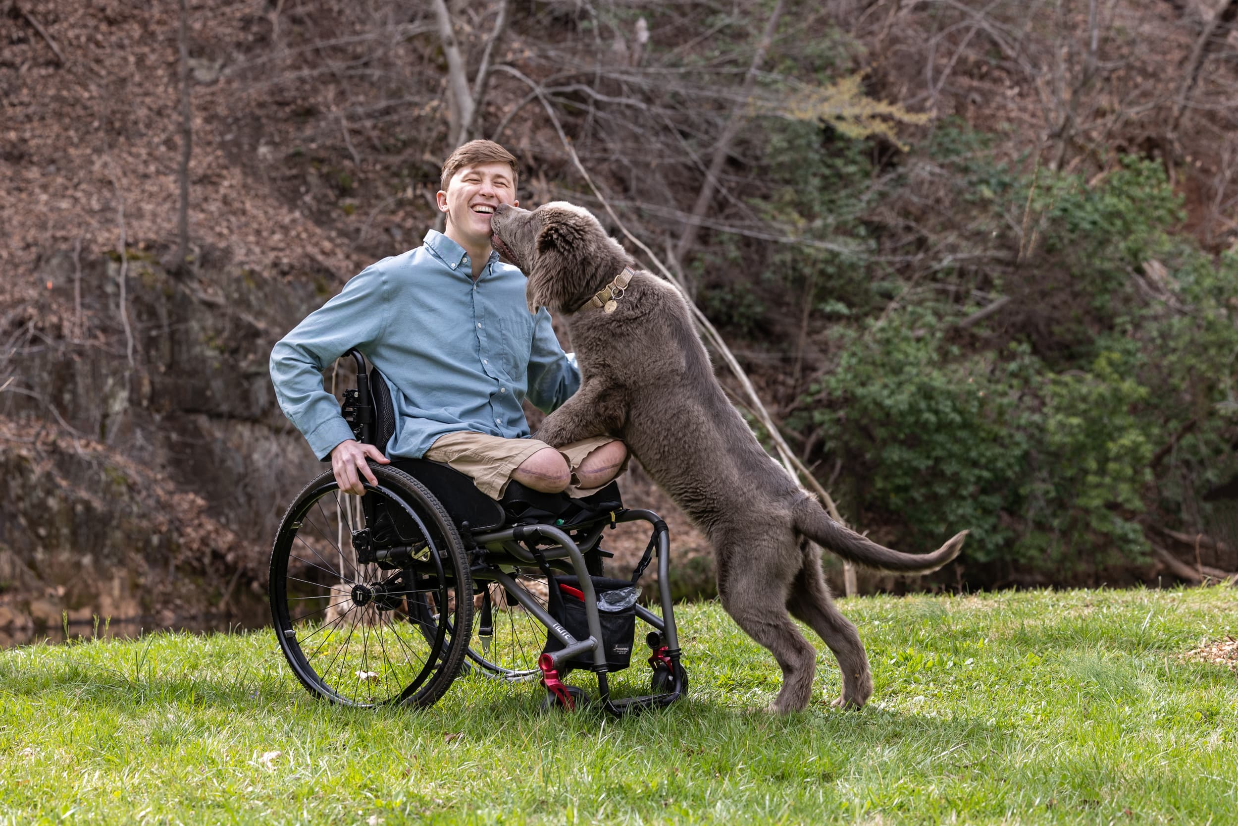 A man in a wheelchair is smiling outside in a grassy area. A large, fluffy brown dog is standing with its front paws on his lap, appearing playful and affectionate. Trees and a rocky hillside are in the background.