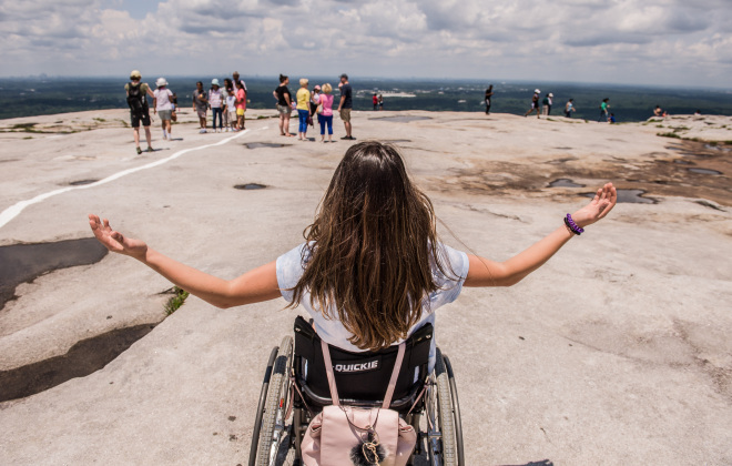 A girl in a wheelchair with outstretched arms, facing a scenic view on a rocky surface. She is surrounded by groups of people in the distance under a partly cloudy sky.