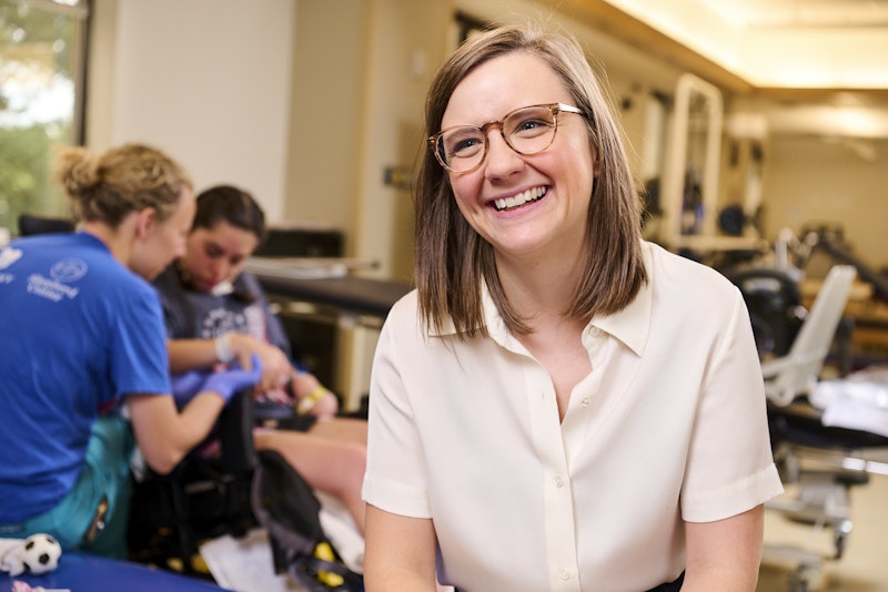 A woman in a white blouse and glasses smiles in the foreground of a physical therapy clinic. In the background, two individuals work with therapy equipment. The setting appears welcoming and professional.