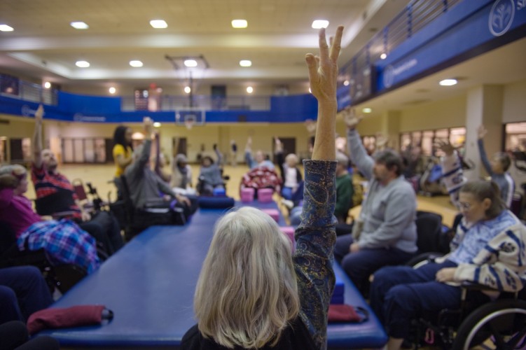 A group of elderly individuals, some in wheelchairs, participate in a seated exercise class in a gym. They are sitting in a circle, raising one arm in the air, engaging actively with the instructor. The gym has blue banners and a basketball court in the background.