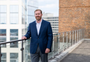 A man with a beard stands on a rooftop walkway, leaning on a glass railing. He is wearing a blue suit jacket over a white shirt, with gray pants. A background of modern buildings and a cloudy sky is visible.