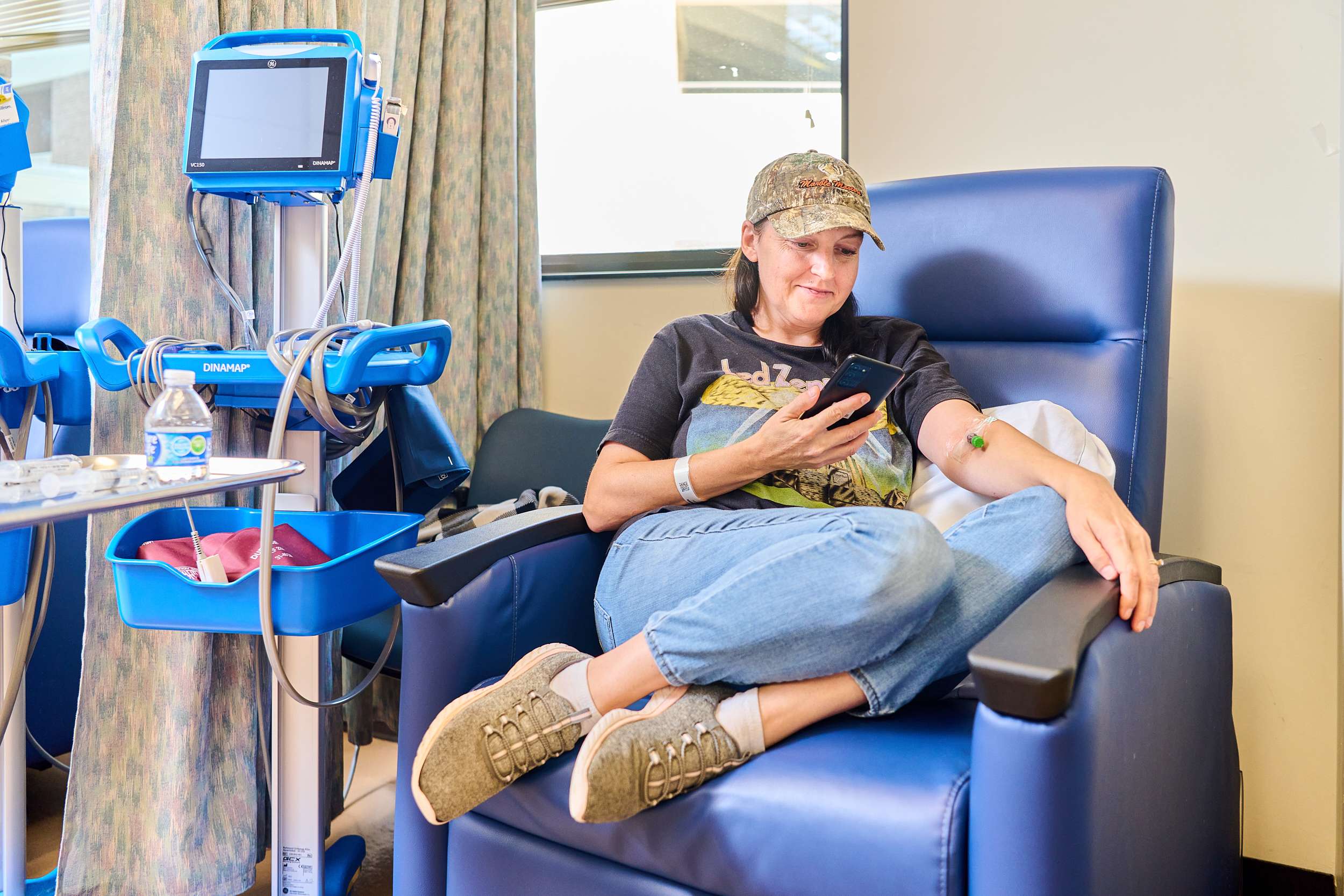 A person sits in a blue recliner at a medical facility, looking at their phone. They're wearing a cap, casual clothes, and have a medical band on one arm. Medical equipment is nearby, and the setting appears calm and relaxed.