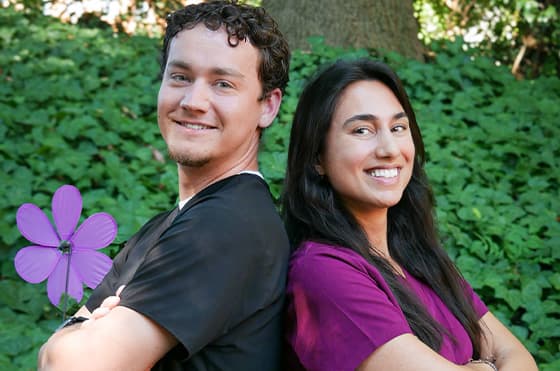A man and woman stand back-to-back, smiling, in front of lush greenery. The man holds a purple pinwheel flower. Both are casually dressed in dark tops.