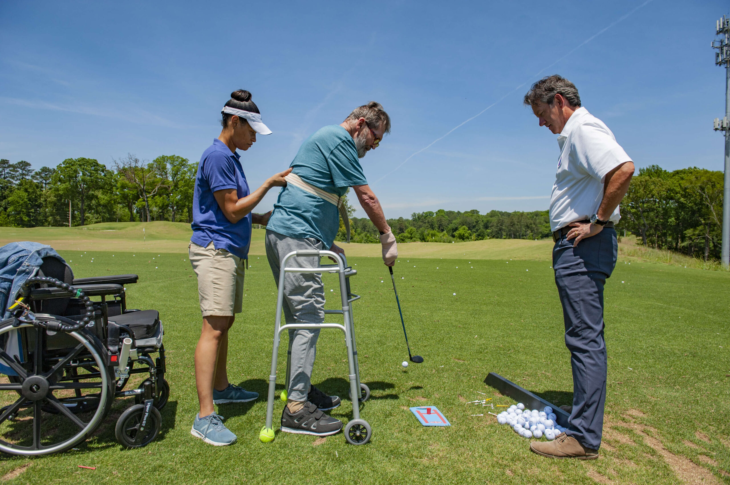 A woman assists an man using a walker as he prepares to hit a golf ball on a driving range. A man in a white shirt stands nearby observing. A wheelchair and basket of golf balls are in the scene. The sky is clear and sunny.