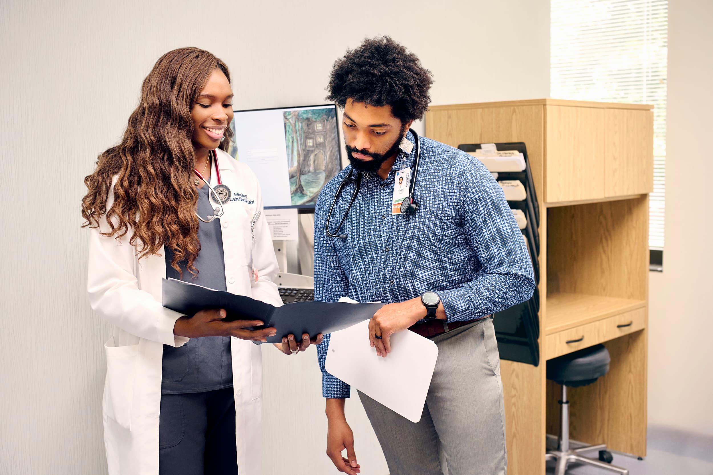 Two healthcare professionals discuss documents in a medical office. The woman, wearing a white coat, holds a folder, while the man, in a blue shirt with a stethoscope, listens intently. Nearby, there is medical equipment and office furniture.