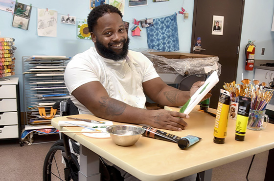 A bearded man in a wheelchair smiles while painting in an art room. He wears a white shirt and has art supplies, including brushes and paint tubes, on the table in front of him. Artwork and photos are displayed on the walls.