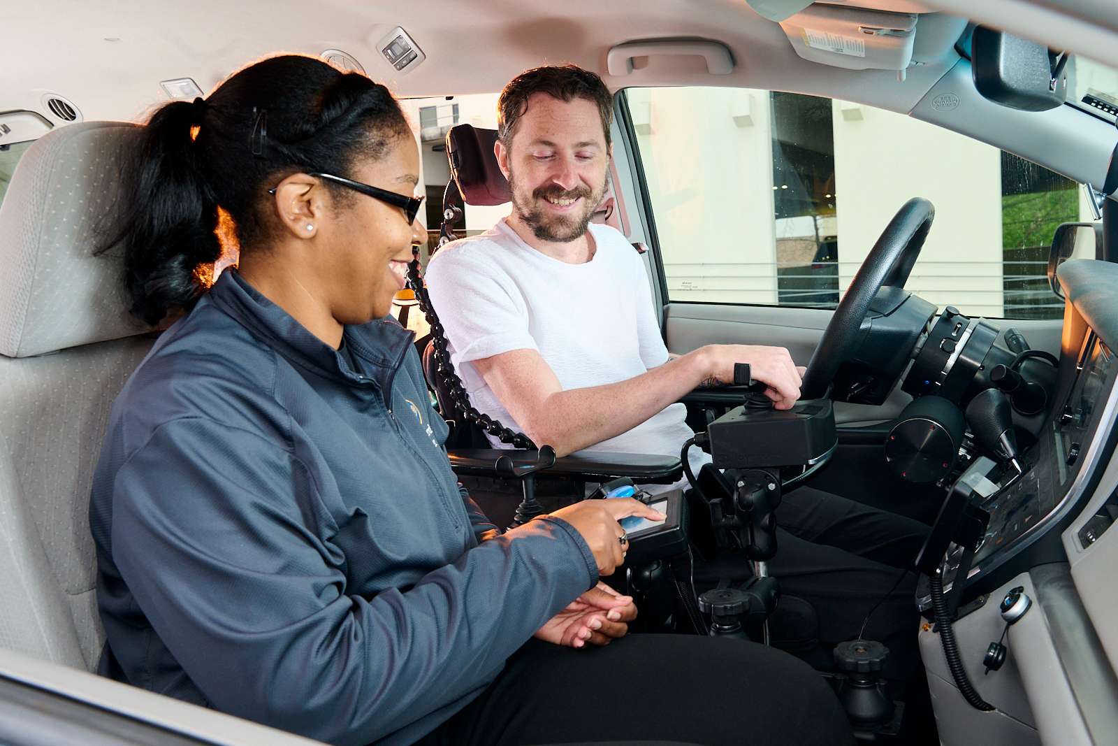 A woman in a gray jacket instructs a man in a wheelchair on how to use adaptive driving controls in a car. The man is smiling and situated next to the driver's seat. The vehicle interior is well-lit.