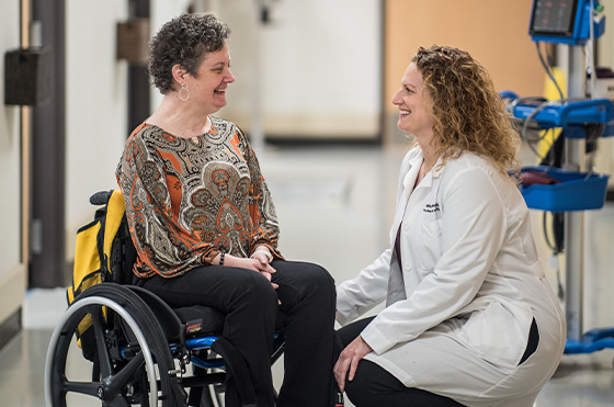 A woman wearing a patterned blouse sits in a wheelchair while smiling and chatting with a healthcare professional kneeling next to her. The healthcare professional is in a white coat and has curly hair. They are in a clinical hallway.