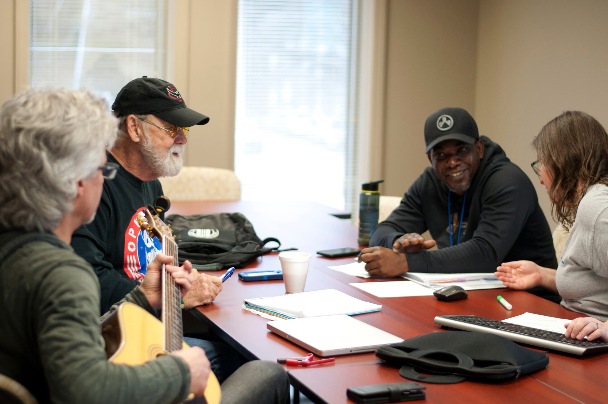 Four people are seated around a wooden table in a conference room, engaged in discussion. One person holds a guitar, and various items including notebooks, a water bottle, a coffee cup, and a keyboard are on the table.