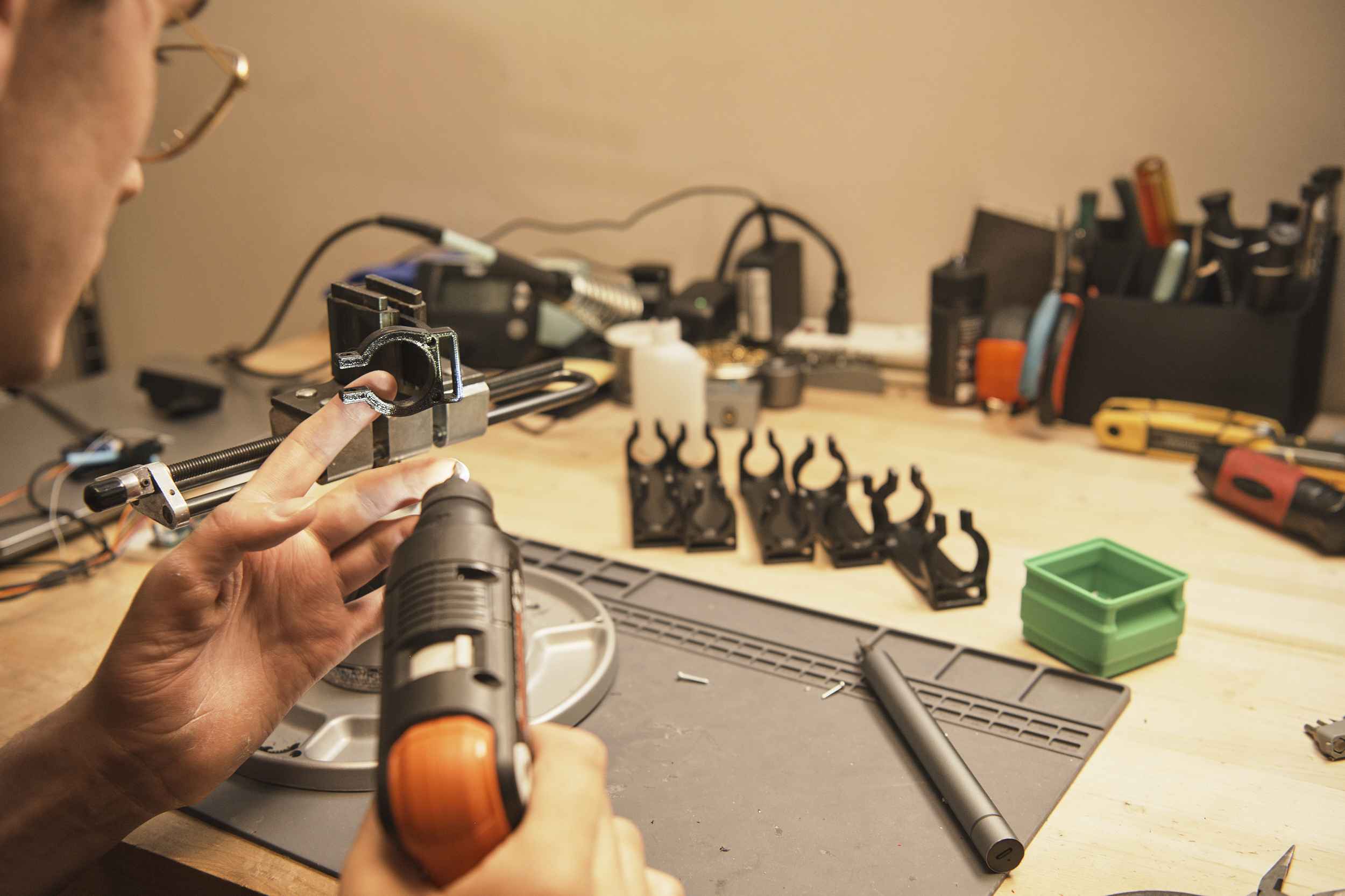 A person uses a tool to examine or work on a small mechanical component, surrounded by various tools and parts on a workbench. The workspace is organized with items like wrenches and a container in the background.