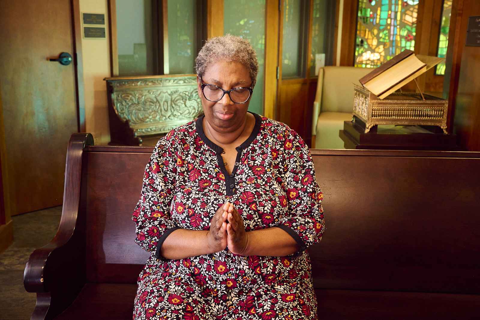 A person with short gray hair and glasses sits on a wooden pew in a dimly lit room with stained glass windows. They are wearing a floral-patterned dress and are holding their hands together in a prayer position.