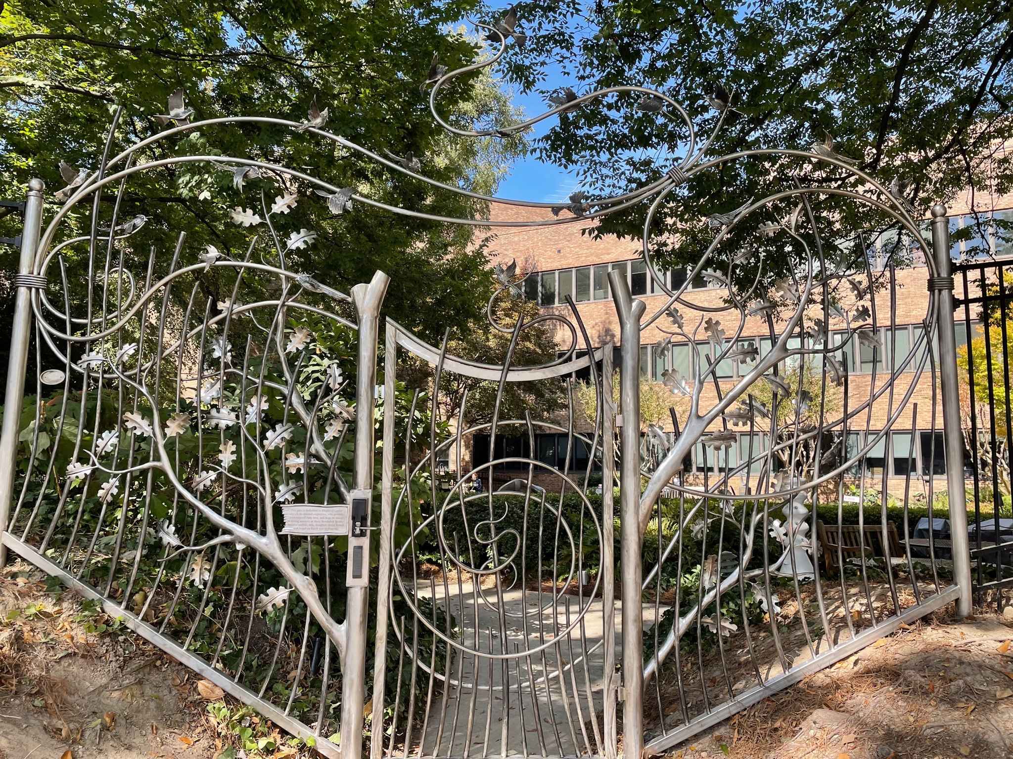 Ornate metal gates featuring intricate floral and vine patterns lead to a pathway surrounded by lush greenery. A brick building with tall windows is visible in the background, under a clear blue sky.