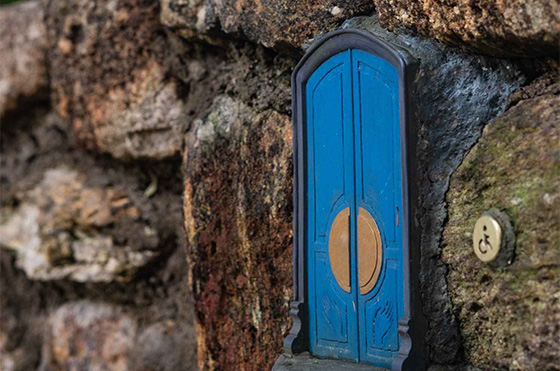 A miniature blue door with a circular golden accent is embedded in a stone wall. Next to the door, there is a small button with a handicapped accessible symbol. The door gives a whimsical, fairy-tale-like appearance to the rugged stone wall backgrounds.