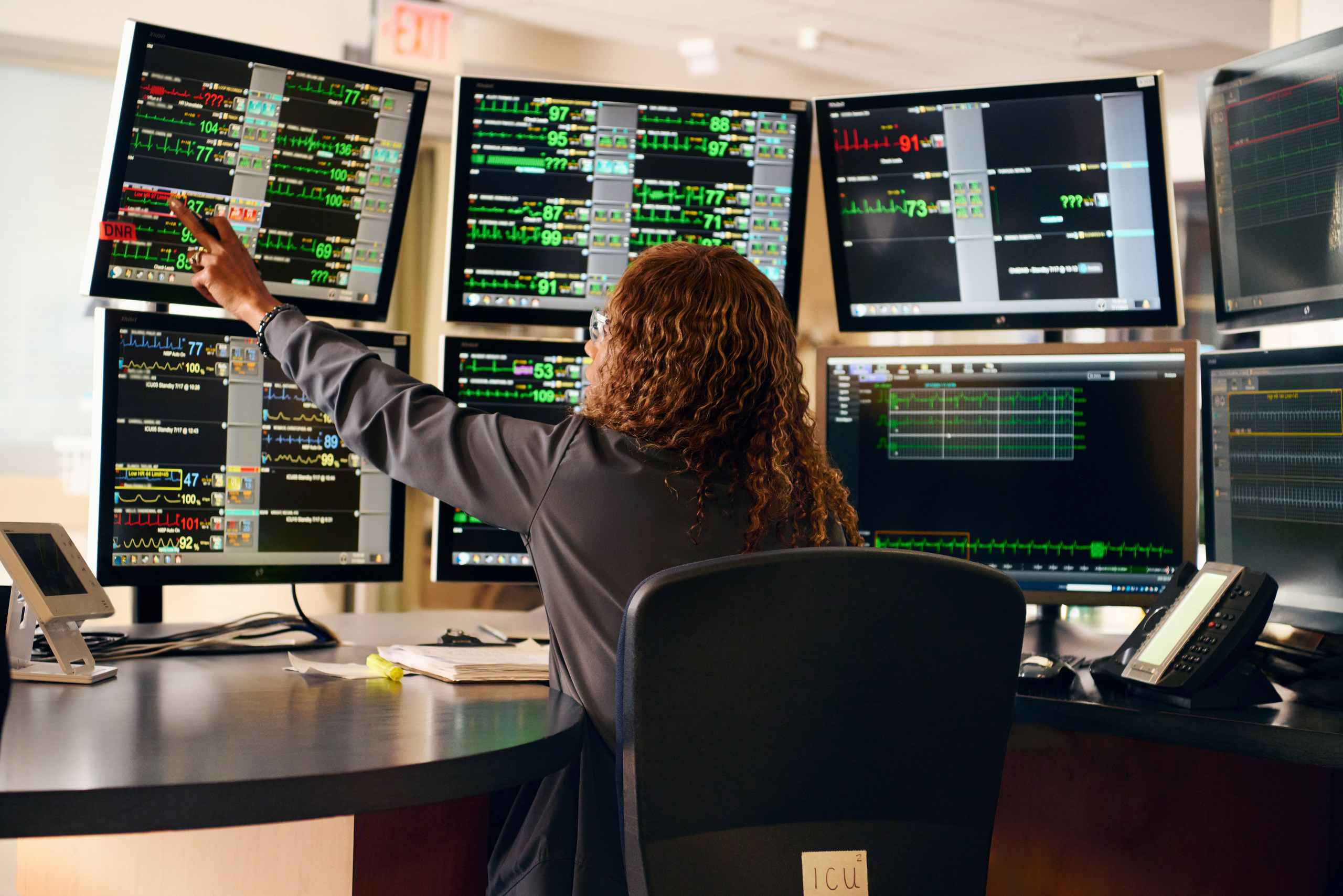 A person sits at a desk surrounded by multiple digital screens displaying medical data, graphs, and vital signs. They point at one of the screens. The environment resembles a hospital monitoring room.