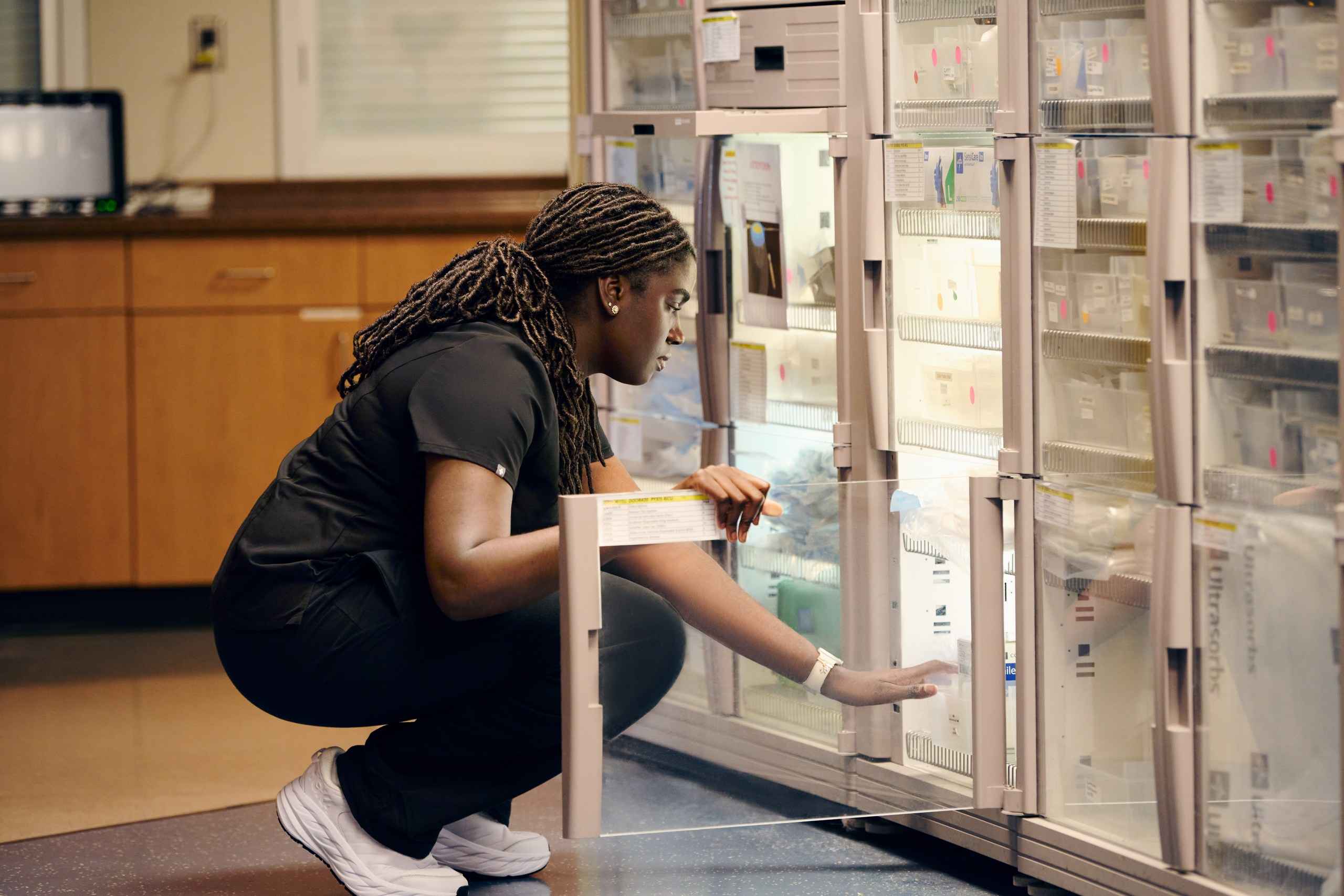 A person wearing scrubs and white sneakers is crouching down in front of a refrigerated storage unit. They are reaching for a medication or item inside. The background shows a room with wooden cabinets and a computer monitor.