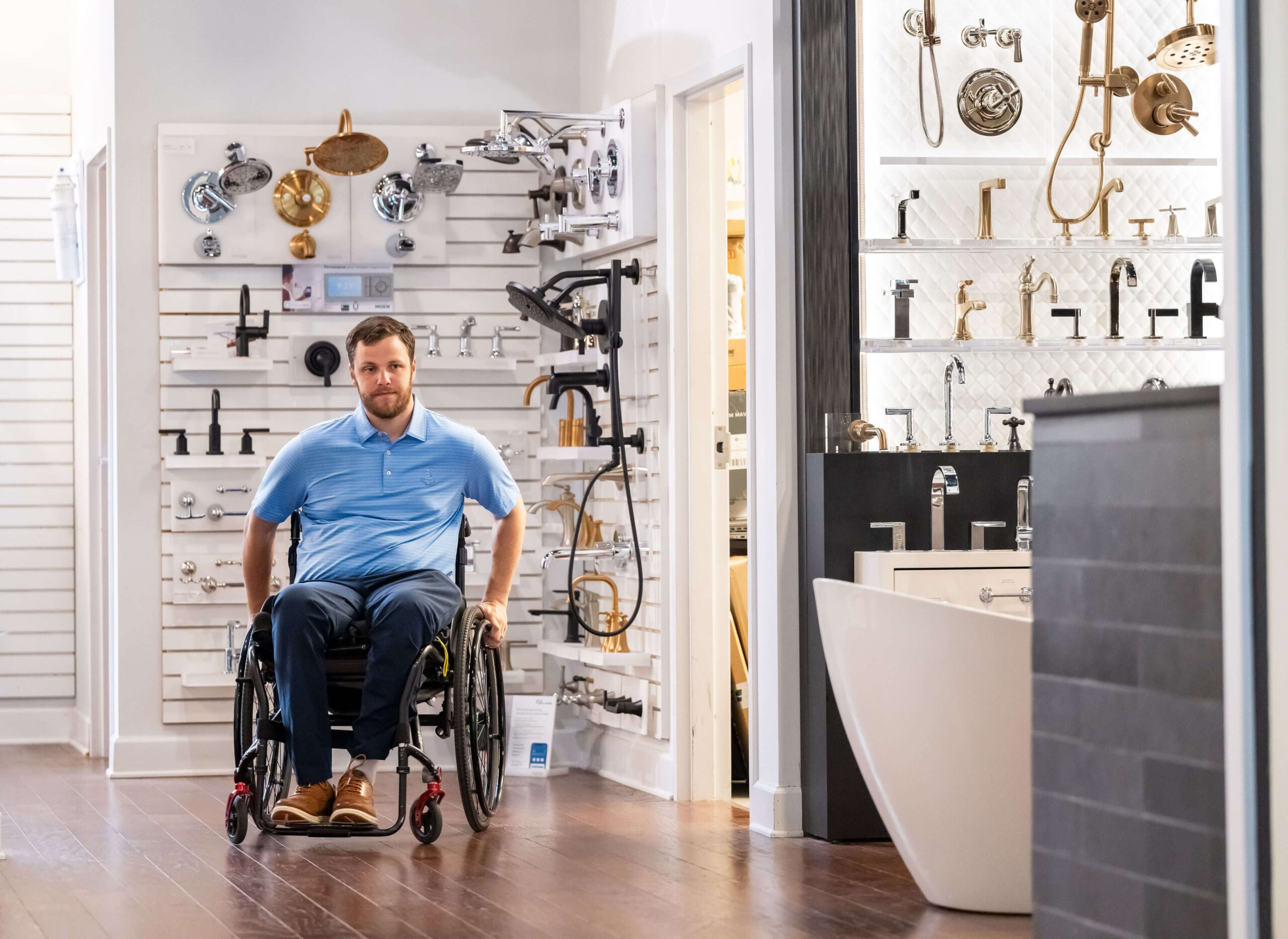 A man in a wheelchair moves through a showroom displaying various bathroom fixtures. There are numerous taps, shower heads, and accessories mounted on display walls around him. The floor is wooden, and there is a white bathtub visible.