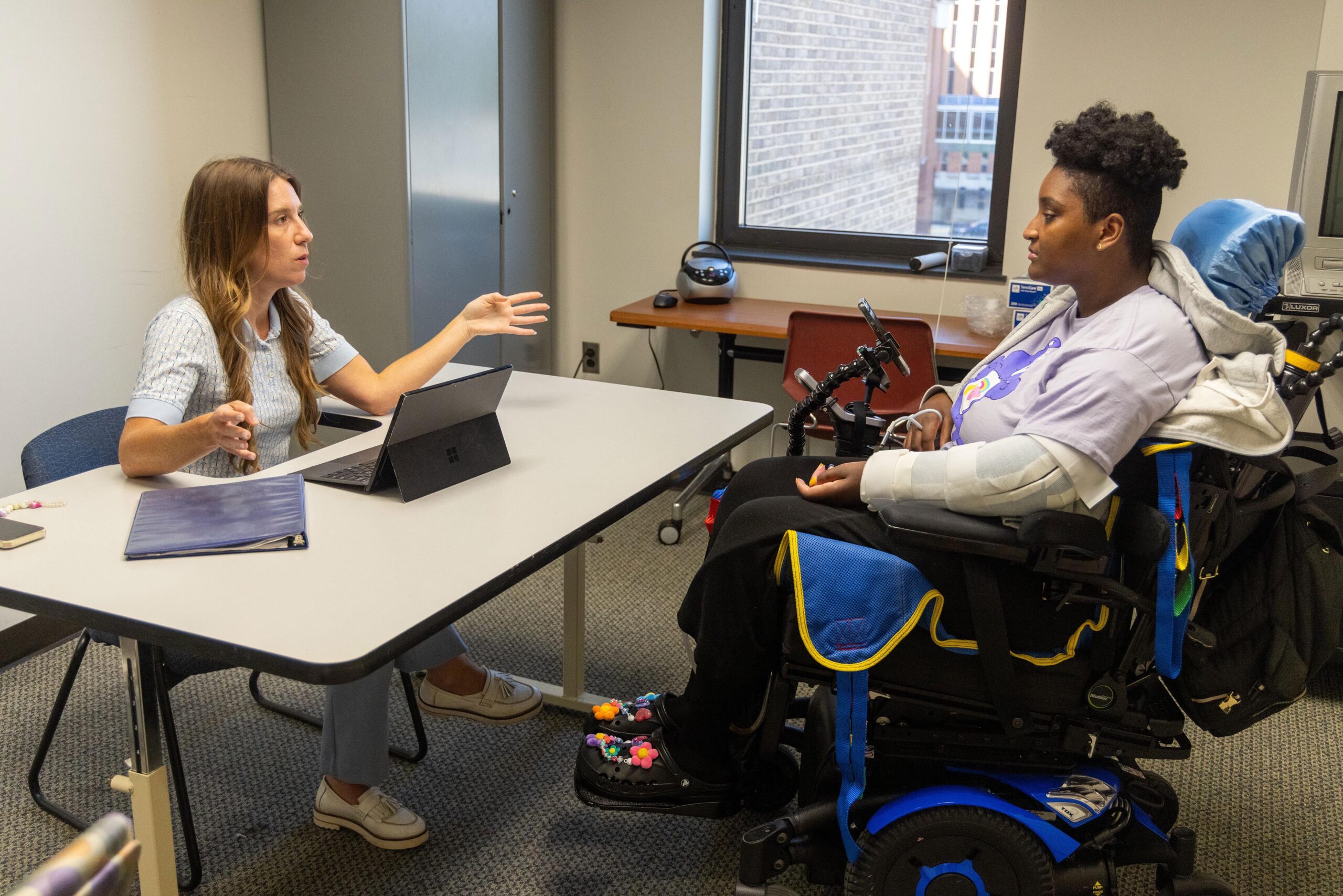 A woman sits at a desk with a laptop, gesturing as she talks to a woman in a motorized wheelchair. They are in an office with large windows.