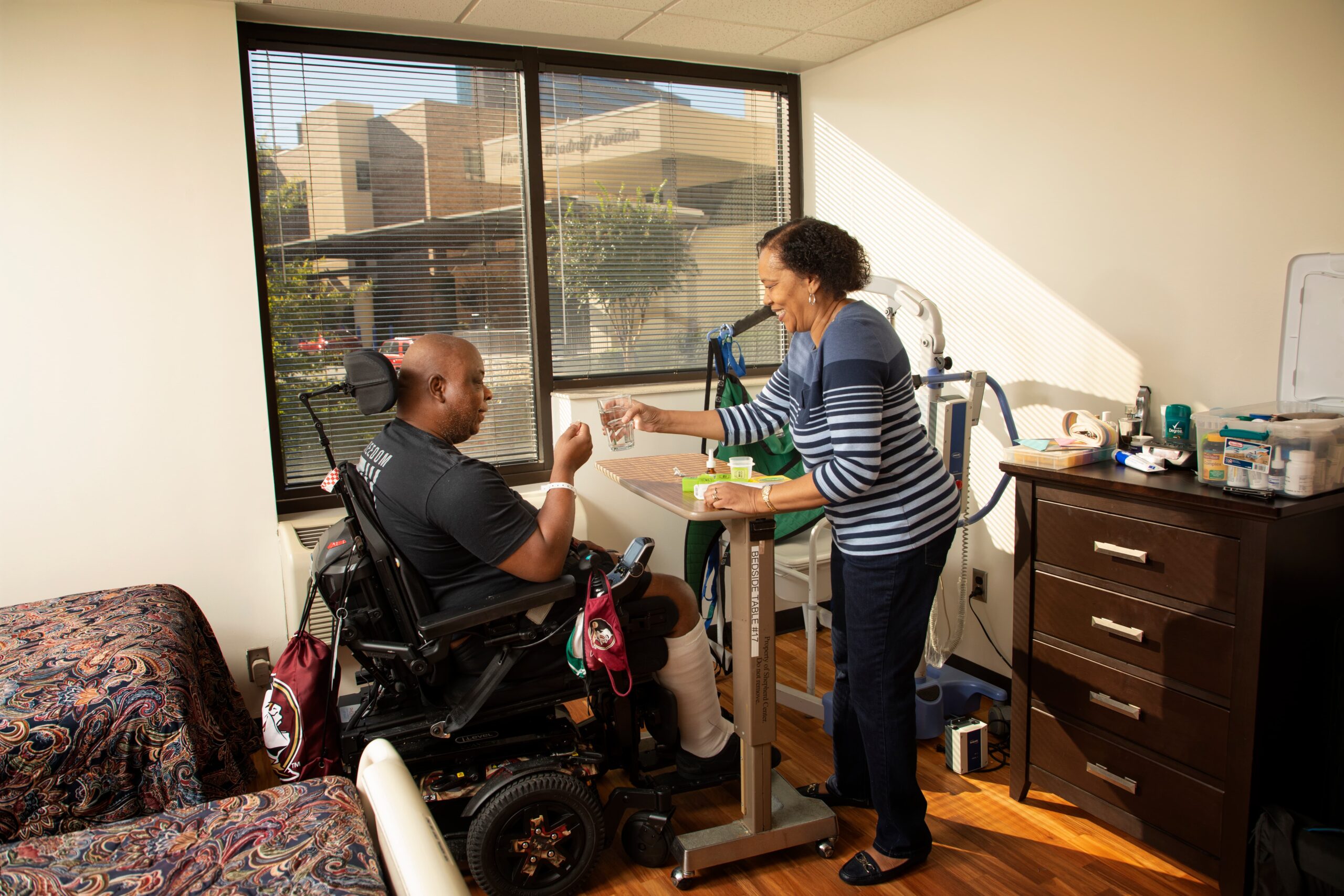 A person in a motorized wheelchair receives a glass of water from a caregiver in a well-lit room. The room has a large window, a bed, bedside table, and medical equipment, creating a comfortable environment.