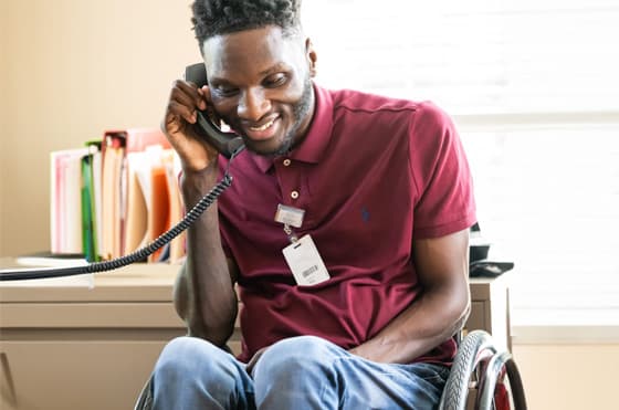 A man wearing a maroon polo shirt, seated in a wheelchair, smiles while talking on a landline phone. He has an identification badge hanging from his neck. Behind him is a desk with various colored files and a window letting in natural light.