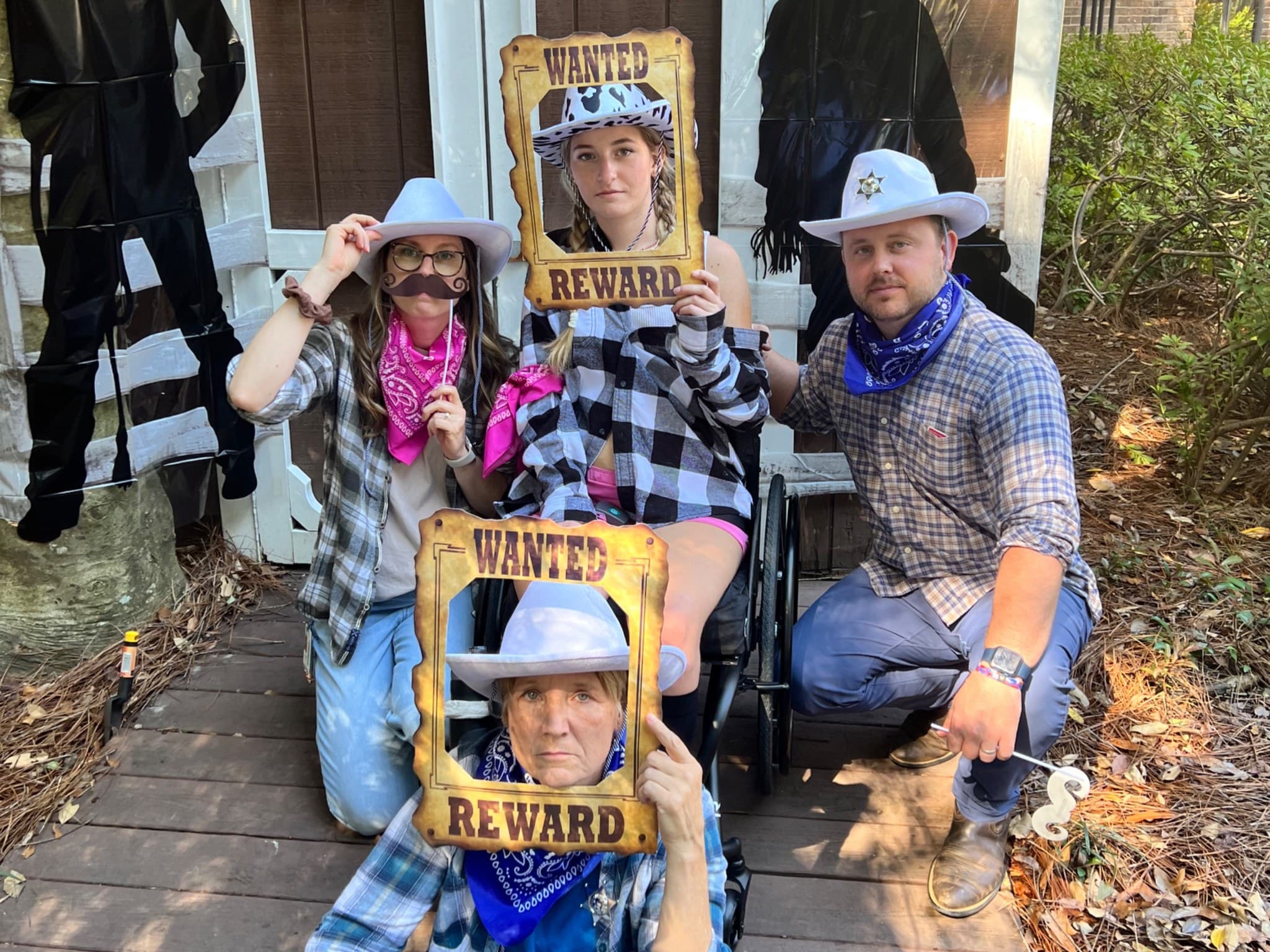 Four people dressed as cowboys and cowgirls pose in front of a wooden structure, holding 