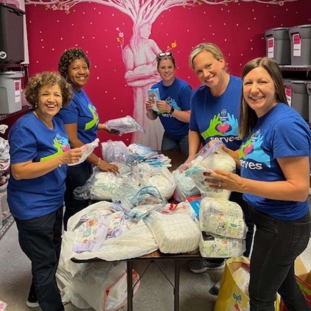 A group of five smiling women in matching blue shirts are sorting and packing diapers and baby supplies in a room with a pink wall featuring a tree mural.