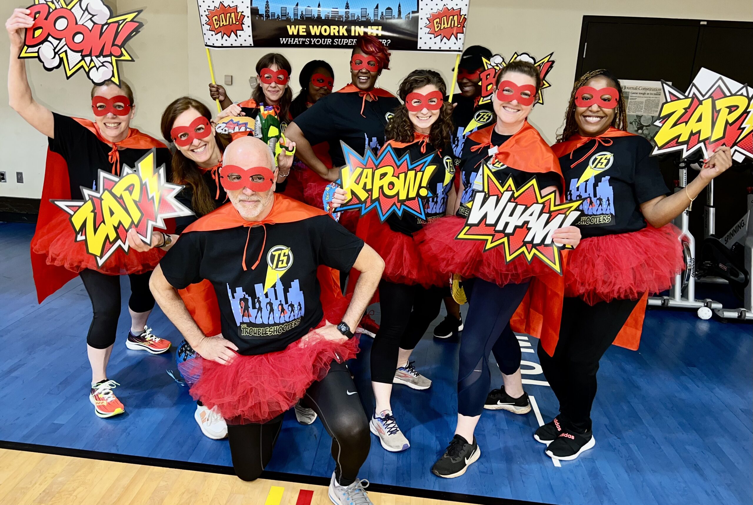 A group of people posing in superhero costumes with red capes, red masks, and tutus. They hold comic-style signs reading 