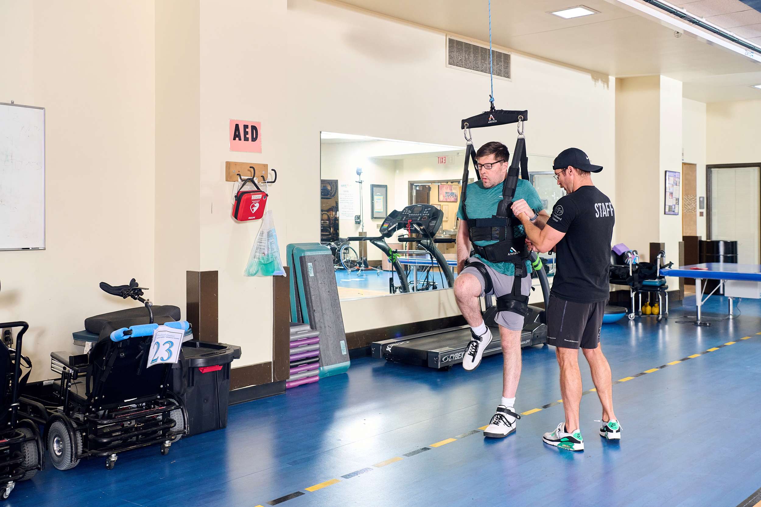 A man in athletic gear assists another man using a harness system for physical therapy or rehabilitation. They are in a spacious room with exercise equipment and a mirror on the wall.