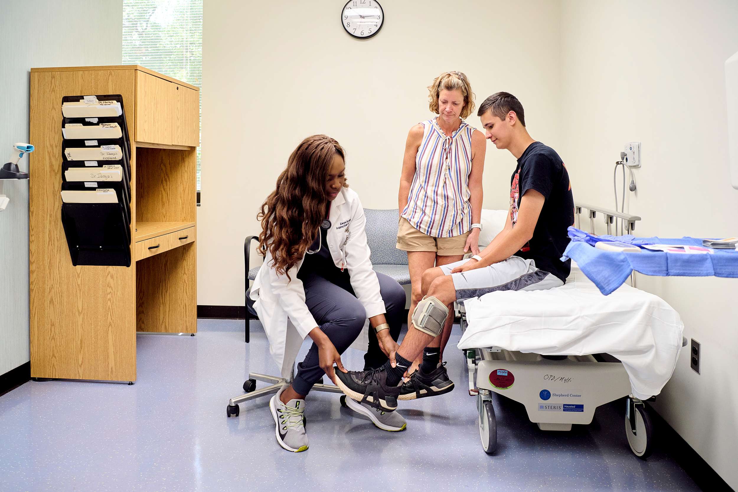 A medical professional helps a seated young man with a knee brace in a clinic room. A woman stands beside them, observing. The room features medical equipment, a clock, and a window with natural light.