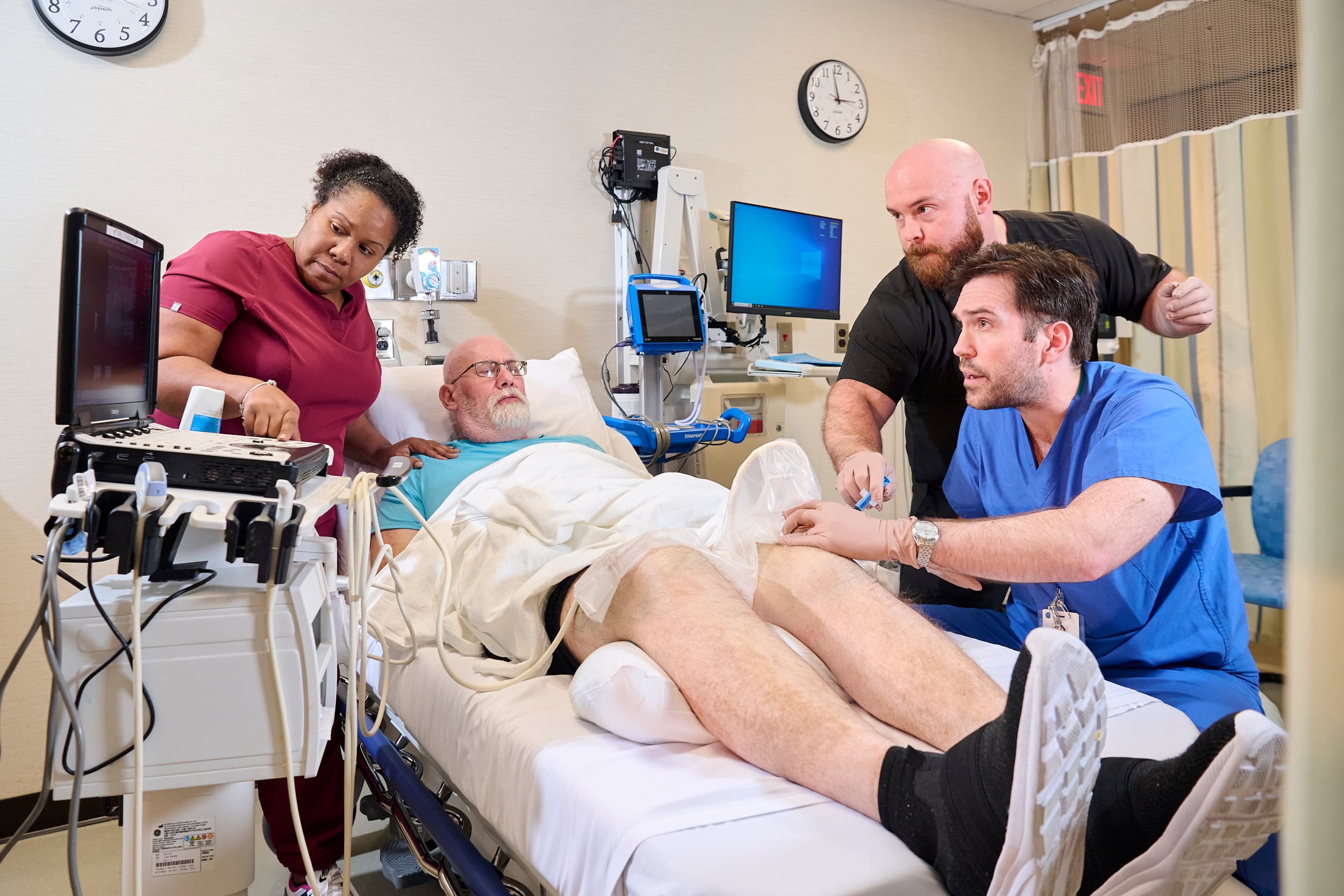 A medical team performs an ultrasound on a patient lying on a hospital bed. A nurse and two doctors are focused on the procedure, using equipment to examine the patient's abdomen. The room is equipped with medical devices and monitors.