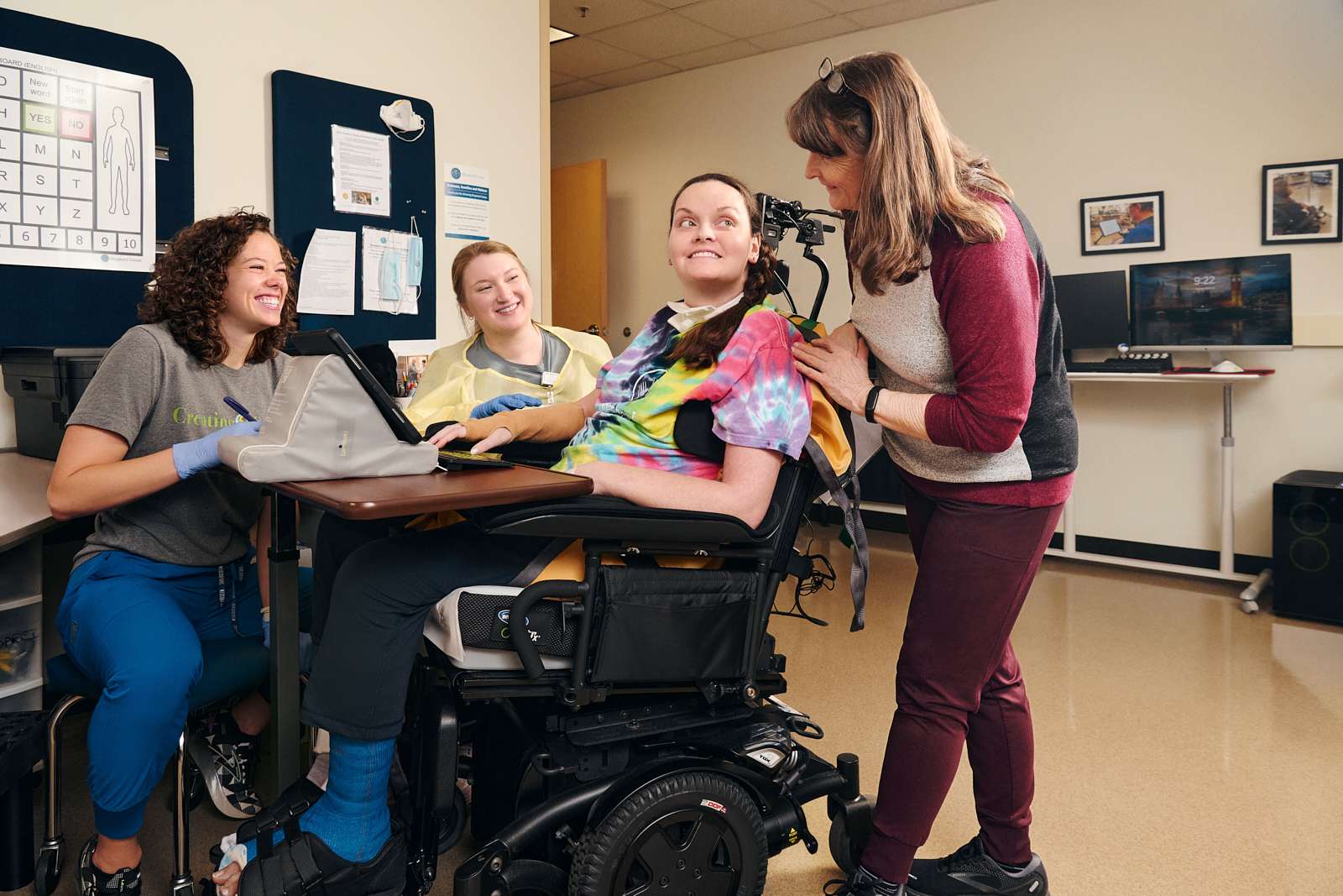 A person in a wheelchair smiles while surrounded by three people. One person kneels beside them, another sits with a laptop, and the third stands, leaning in with a hand on their shoulder. The room has a chart and computer monitors in the background.