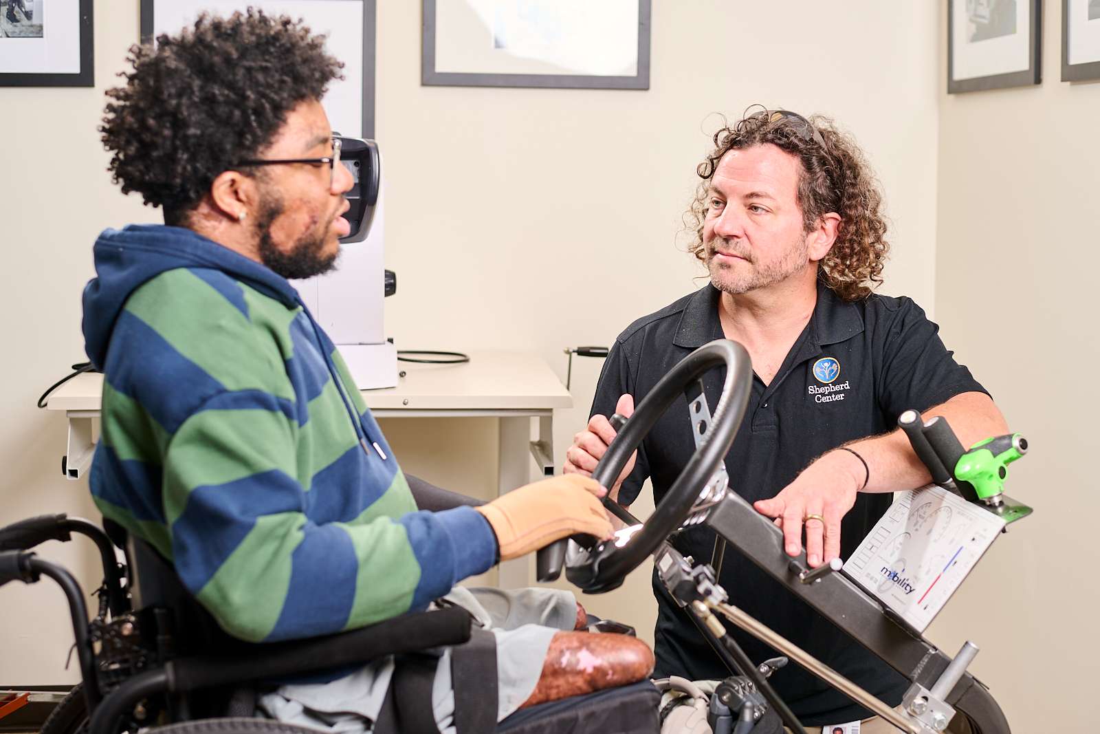 A person in a striped hoodie sits in a wheelchair, speaking with another person with curly hair, wearing a black polo shirt. They are in an indoor setting with framed pictures on the walls in the background.