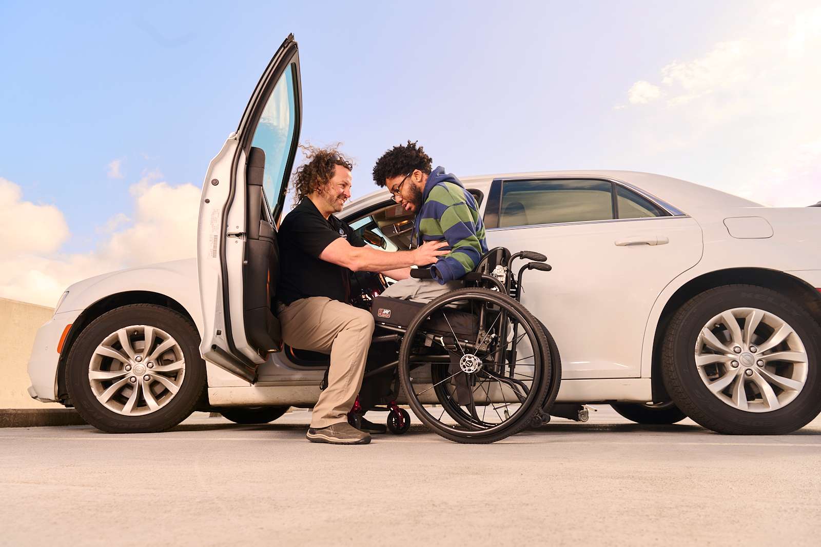 A man in a wheelchair is being assisted by another man to transfer from a car on a sunny day. The car door is open, and they are positioned next to the vehicle. Both are smiling, and the sky is clear.