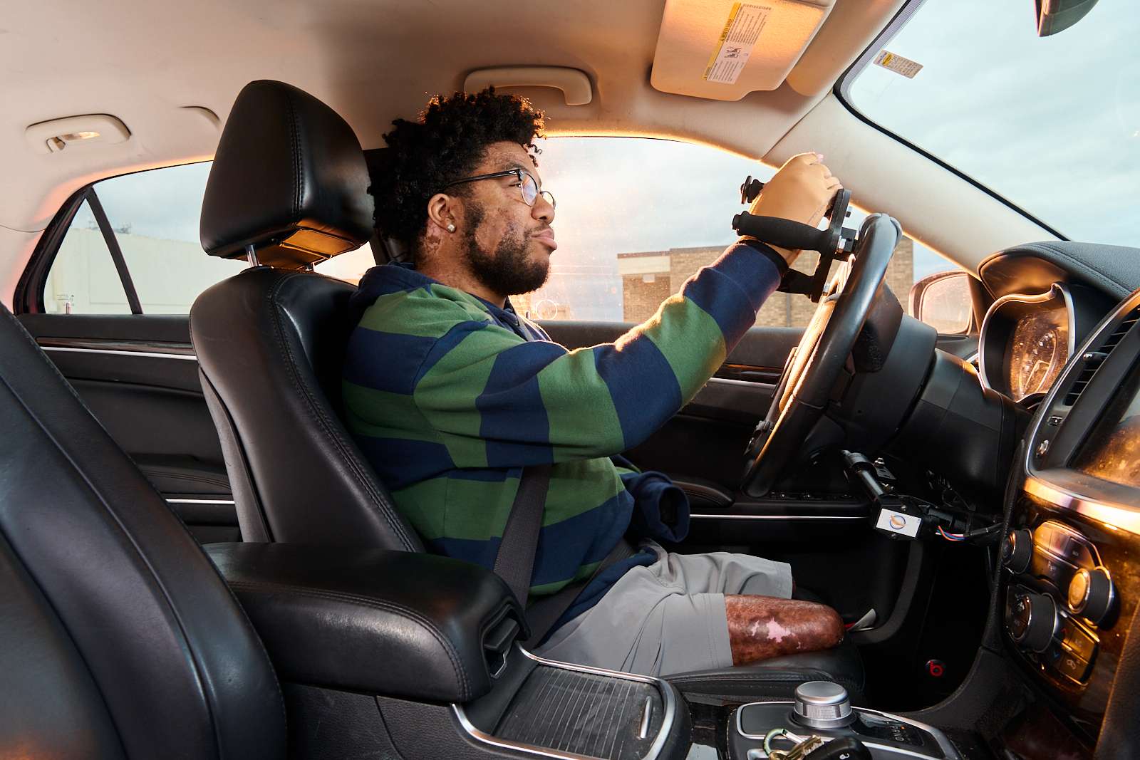 A person with a prosthetic leg drives a car. They are wearing glasses and a striped shirt, focused on the road ahead. The sun casts a warm glow inside the vehicle through the windshield.