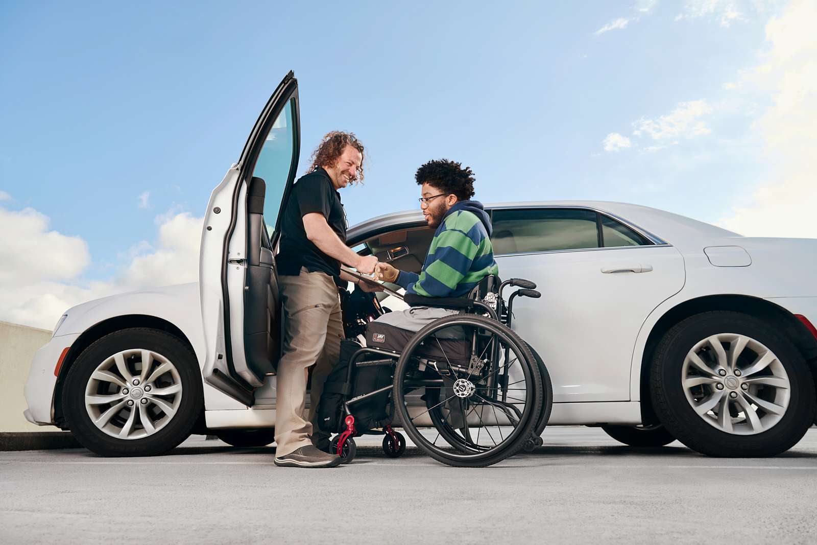 A man in a wheelchair is assisted by another man at an open car door. The car is white, and they are outdoors under a blue sky with clouds.