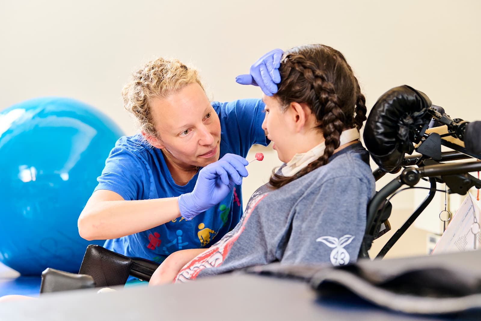 A speech therapist in a blue shirt and purple gloves is interacting with a young person in a wheelchair. They are holding a lollipop near the person's mouth. A large blue exercise ball is in the background.