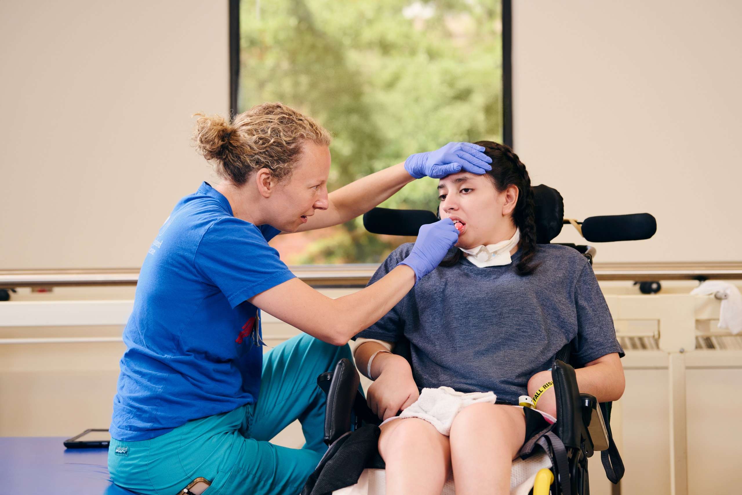 A caregiver in blue scrubs and gloves is examining a young person seated in a wheelchair, wearing a tracheostomy tube. They are in a bright room with a window showing green foliage outside.