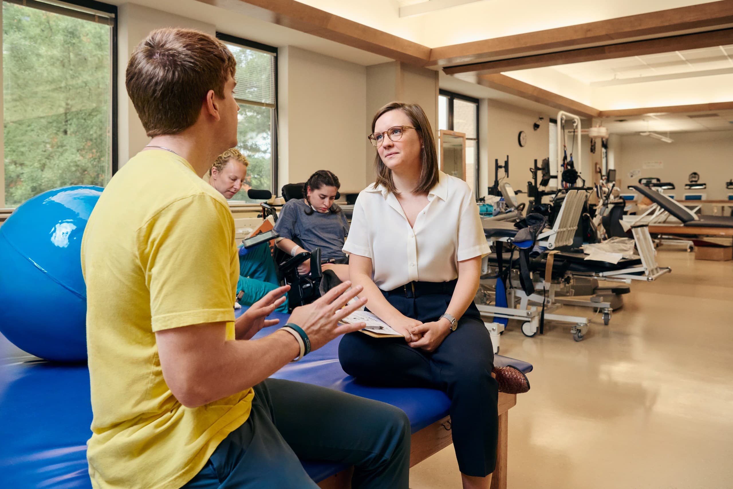 A man in a yellow shirt talks to a woman in a white blouse, seated on a blue therapy table in a rehabilitation center. In the background, another woman in a wheelchair and a person on therapy equipment can be seen.