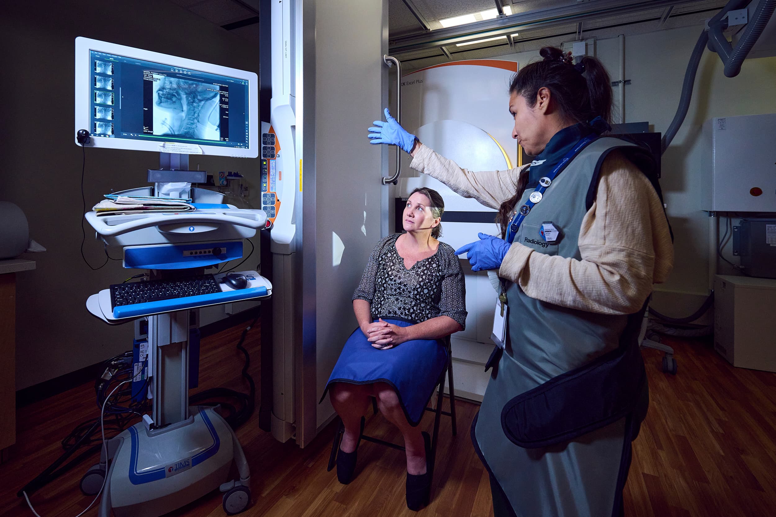 A medical professional assists a patient in a radiology room. The patient is seated next to a large imaging machine, while the professional operates a computer displaying the patient's scans. Both are wearing protective gear.