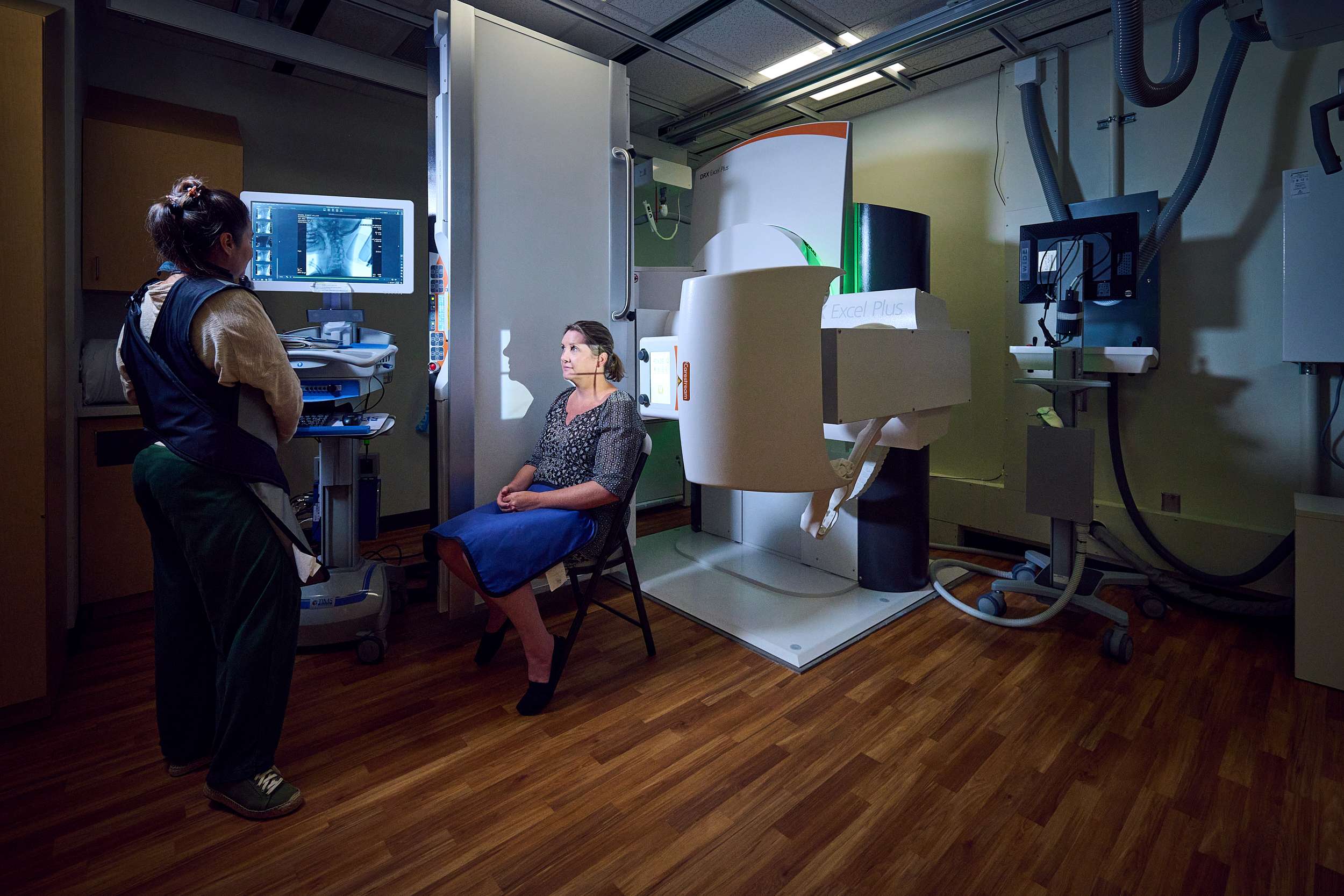 A woman sits on a chair inside a medical imaging room with advanced equipment surrounding her. A technician stands nearby, reviewing images on a screen. The room has a wood-patterned floor and dim lighting.