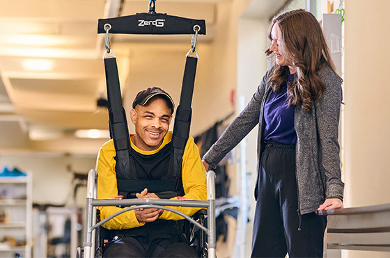 A man in a yellow shirt smiles while seated in a harness system, which is being adjusted by two women. They are in a rehabilitation facility with bright lighting and exercise equipment in the background.