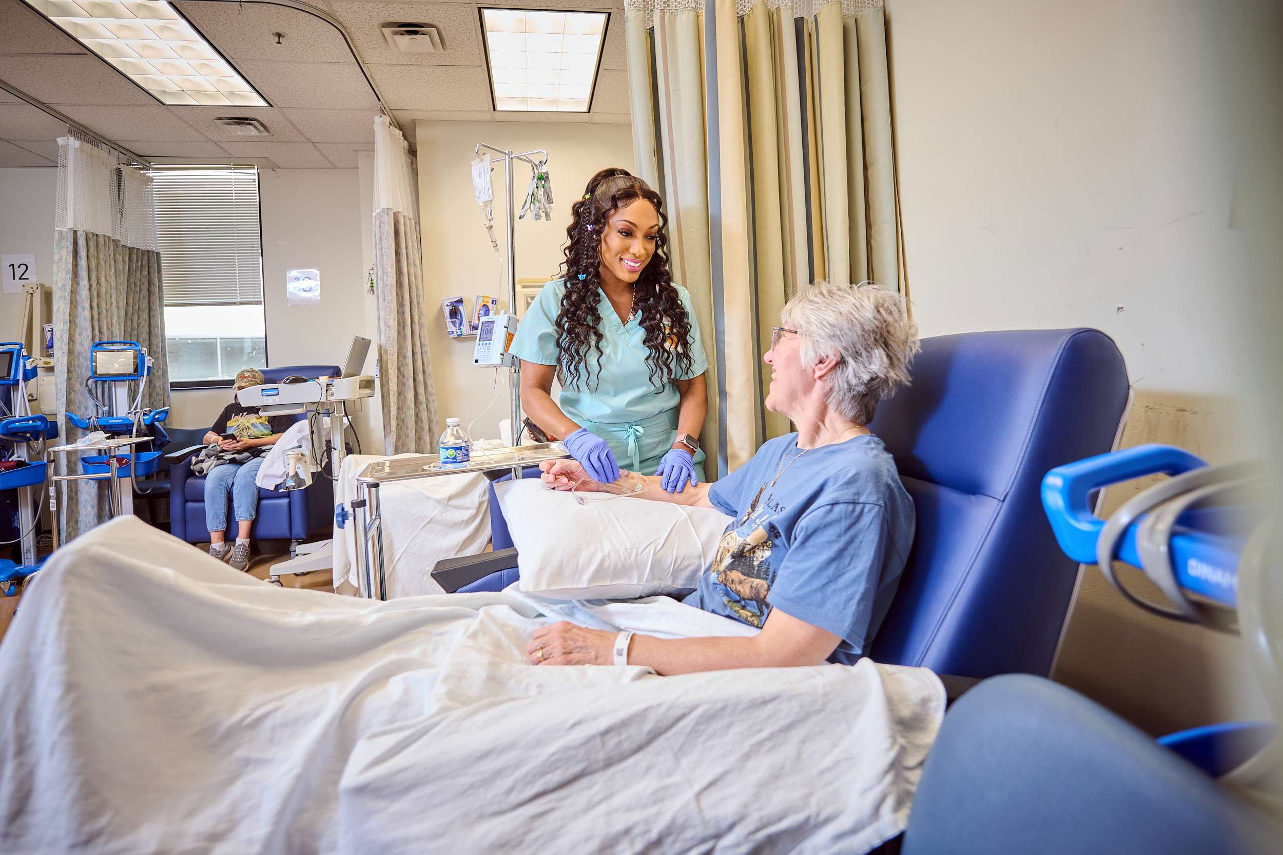 A healthcare worker in scrubs smiles while talking to a patient in a hospital chair. The patient, wearing glasses and a blue shirt, smiles back. Medical equipment is visible in the background.