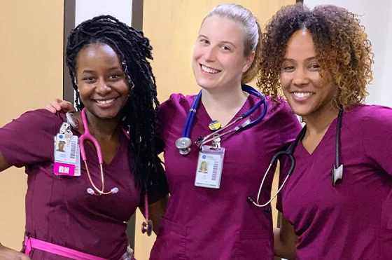 Three healthcare professionals wearing purple scrubs and stethoscopes smile together in a medical setting. They each have identification badges, and the background shows light-colored doors.