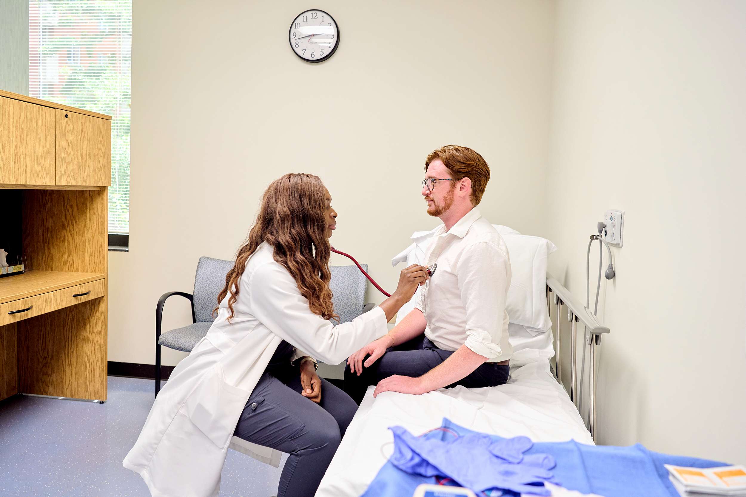 A healthcare professional listens to a patient's heartbeat with a stethoscope in a medical examination room. The patient is seated on an examination table. A clock, cabinet, and chair are in the background. Medical gloves are on the table.