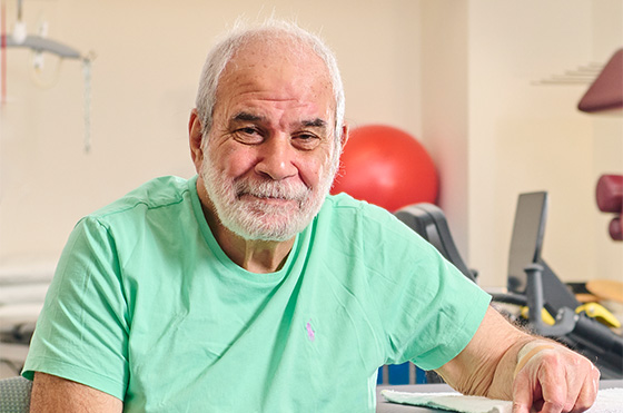 Elderly man with a beard wearing a light green shirt sits on the edge of a medical examination table, smiling. The room has medical equipment visible in the background.