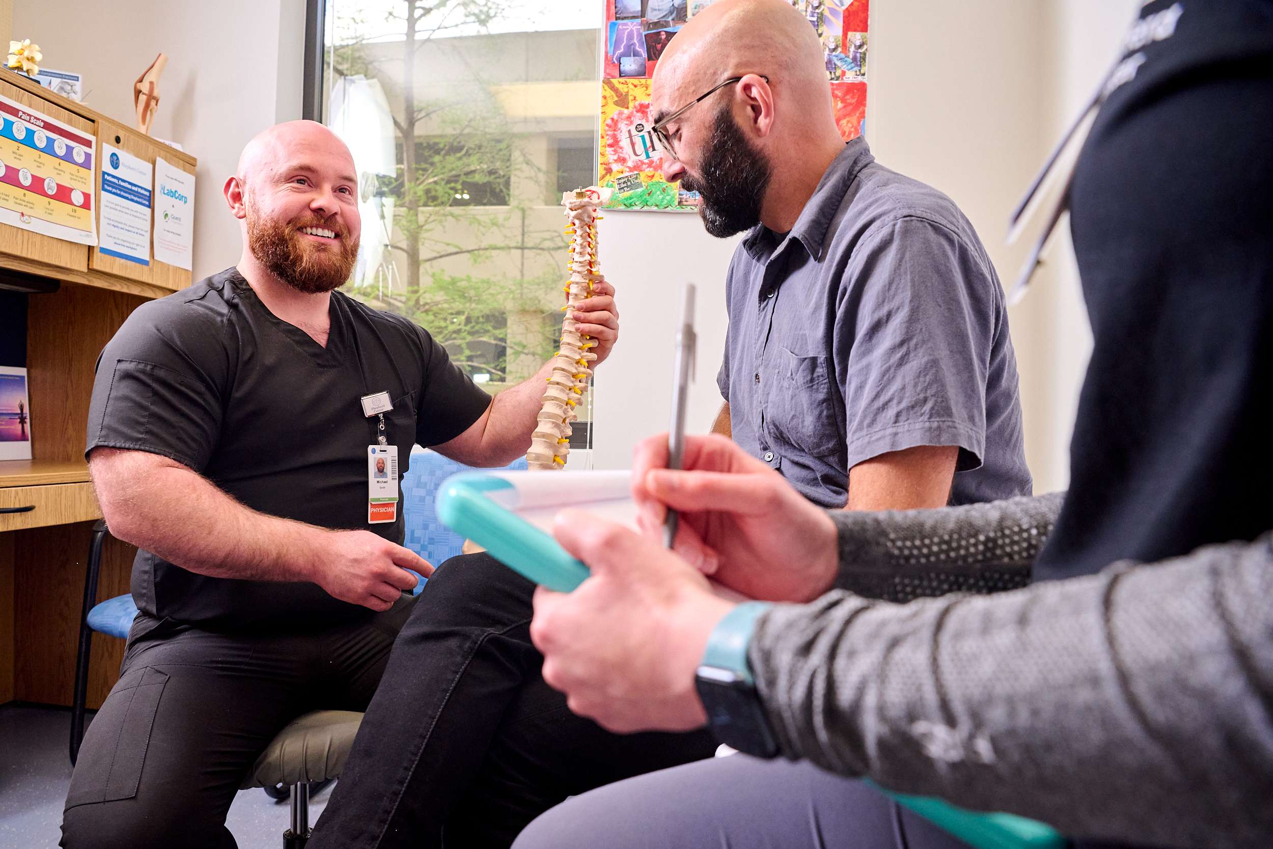 A medical professional in a black uniform shows a spine model to a seated man with glasses and a beard. Another person is taking notes on a clipboard. The setting appears to be a consultation room.