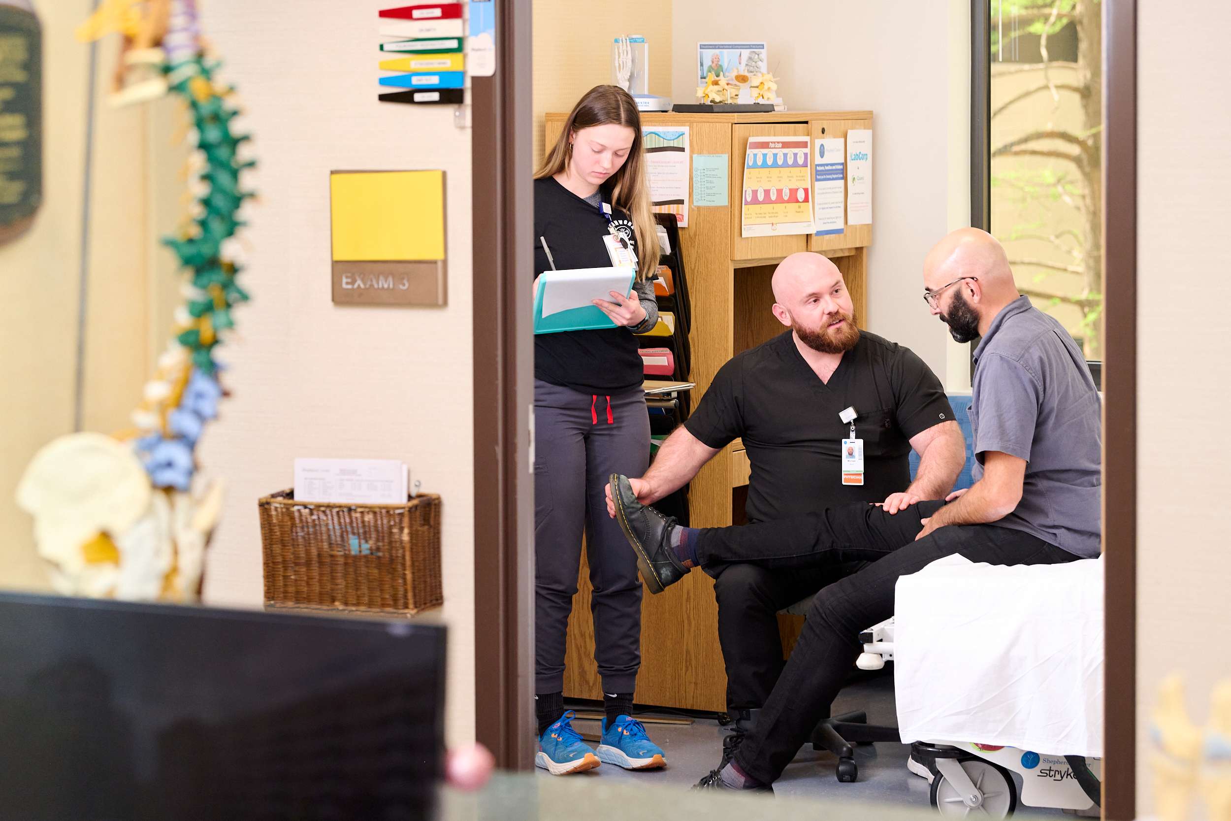 A healthcare professional examines a patient's foot while seated on an exam table. Another person stands nearby taking notes. The room is equipped with medical charts and anatomical models.