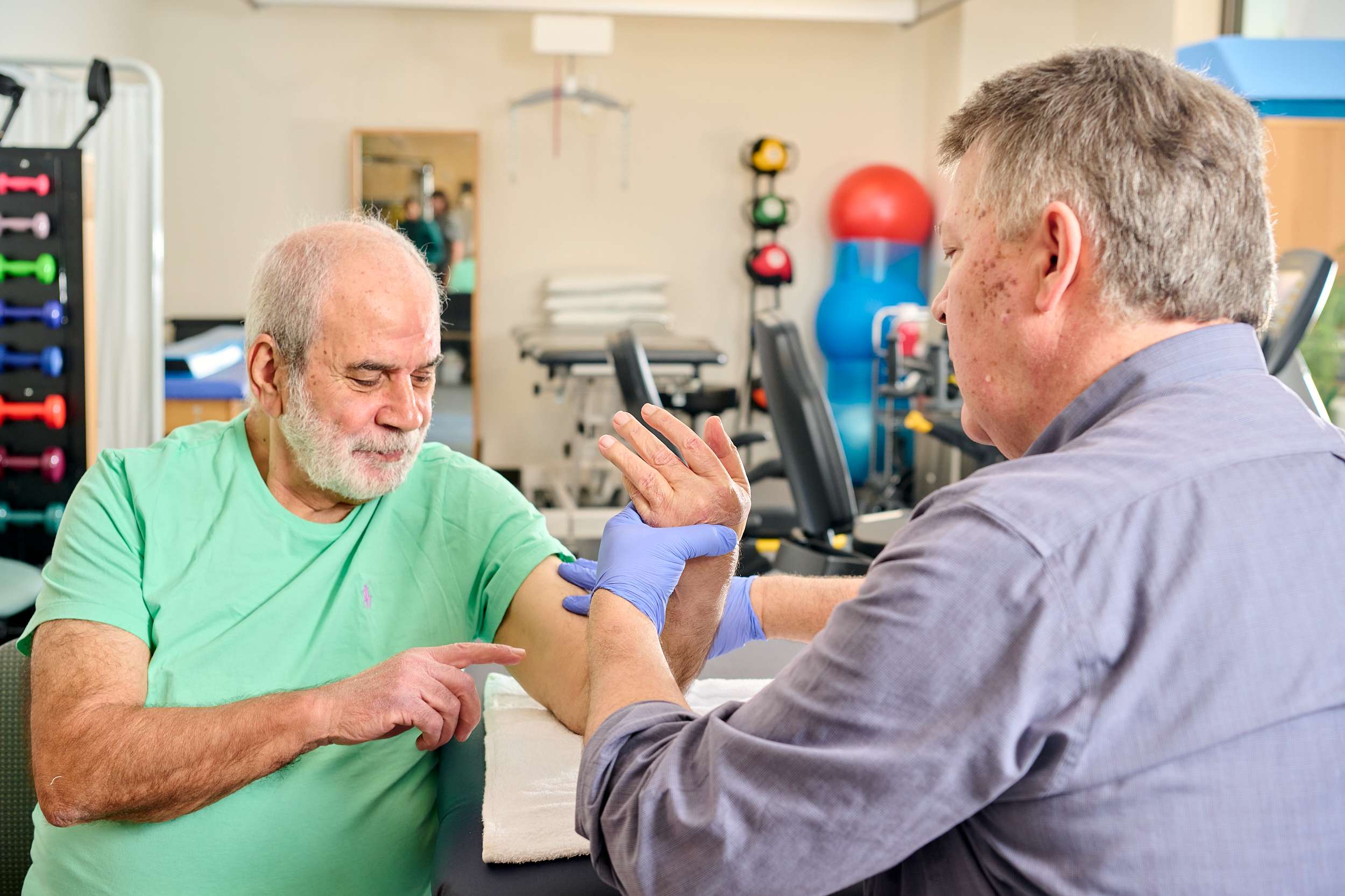 A physical therapist in a gray shirt and gloves assists an older man in a green shirt with arm exercises, aimed at alleviating chronic pain, in a rehabilitation center. Gym equipment and colorful exercise balls add vibrancy to the room's backdrop.