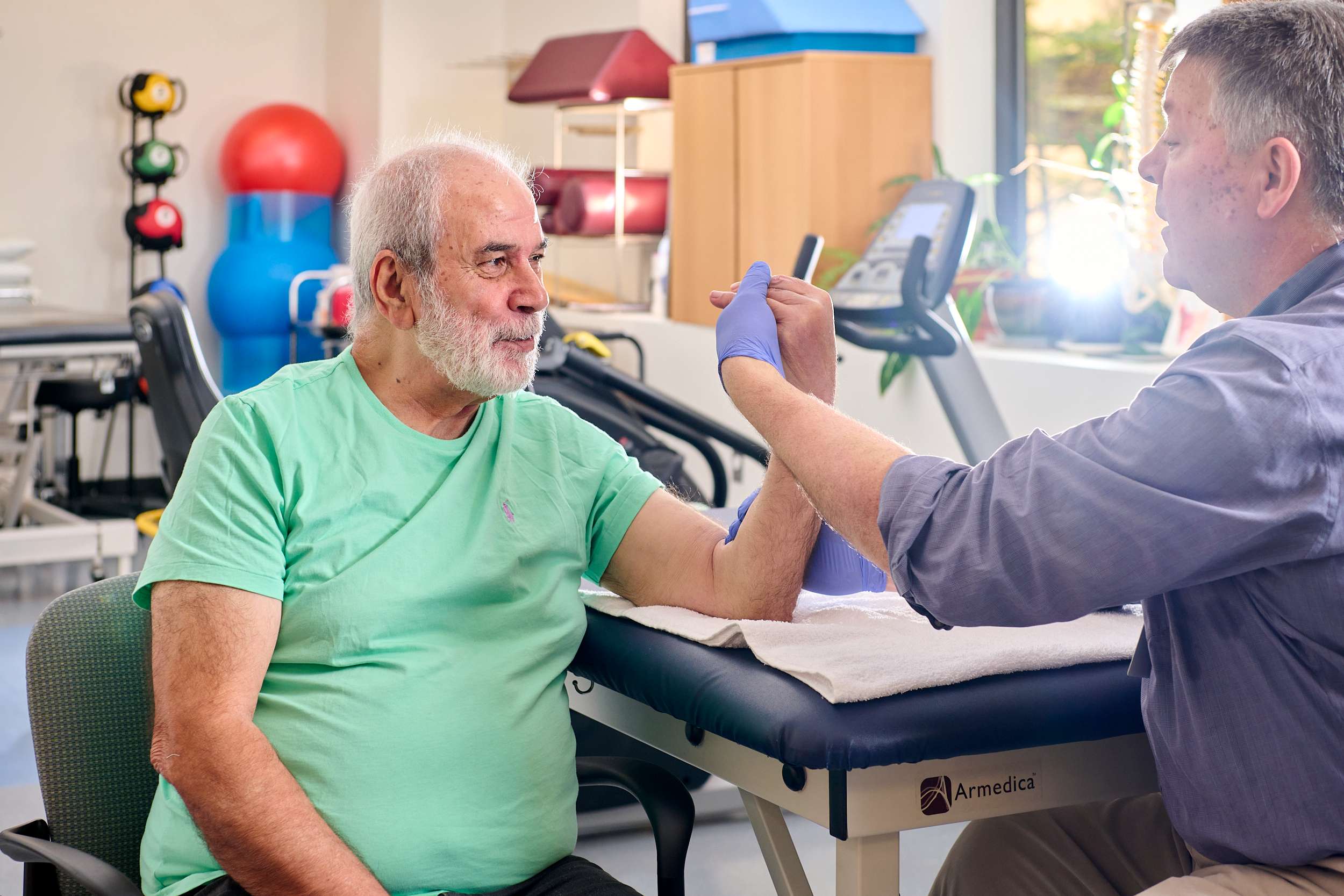 Elderly man in a green shirt receives physical therapy in a clinic. Therapist holds and extends his arm. Exercise equipment is visible in the background.