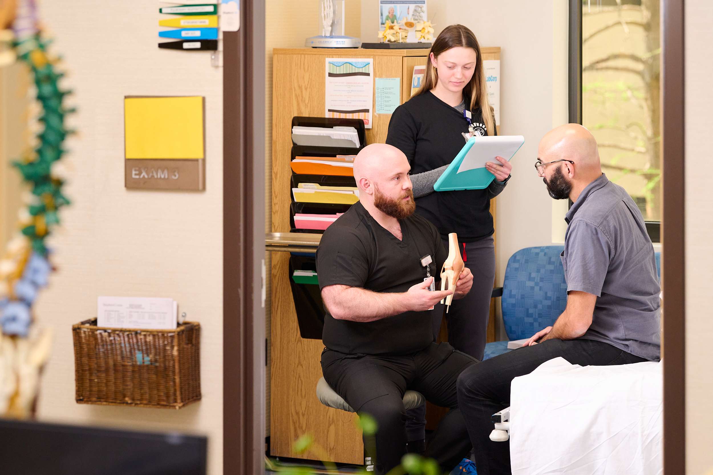 A healthcare professional in black scrubs sits, holding a model spine, explaining to a patient seated on a bed. Another professional stands nearby, taking notes on a clipboard. The room has medical supplies and decor.